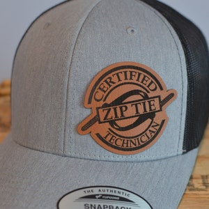 May include: A gray and black baseball cap with a brown leather patch that says "Certified Zip Tie Technician".