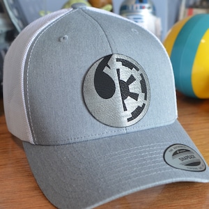 May include: A gray and white baseball cap with a black and silver Star Wars Rebel Alliance symbol patch. The cap has a mesh back and a snap closure. The text "The Authentic Snapback" is printed on the back of the cap.