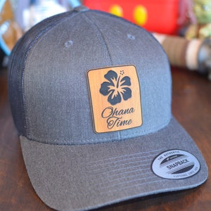 May include: A gray and black trucker hat with a wooden patch that says "Ohana Time" and features a black hibiscus flower. The hat has a black mesh back and a gray front.