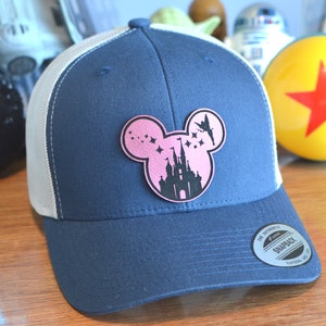 May include: A blue and white trucker hat with a pink Mickey Mouse silhouette featuring Cinderella's castle and Tinkerbell flying above. The hat has a black embroidered patch that says "The Authentic Snapback" with the text "Too Good To Be True" underneath.