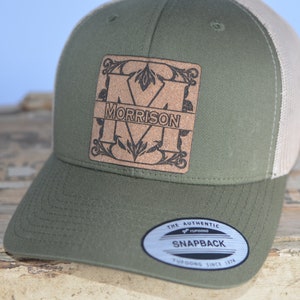 May include: A green and beige trucker hat with a brown leather patch featuring the letter "M" and the name "MORRISON" in a decorative font. The hat has a mesh back and a snap closure.