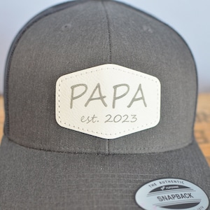May include: A black and white trucker hat with a white leather patch that says "PAPA est. 2023".