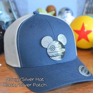May include: Navy blue and white trucker hat with a silver patch featuring a Death Star design in the shape of Mickey Mouse ears. The hat has a curved brim and a Snapback closure.