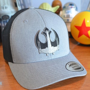 May include: A gray and black baseball cap with a black and silver Rebel Alliance symbol embroidered on the front. The cap has a mesh back and a curved brim. The cap has a "The Authentic" Snapback tag on the back.