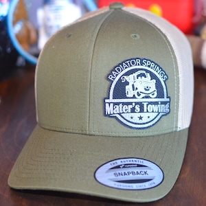 May include: Green and white trucker hat with a black and white patch that says "Radiator Springs Mater's Towing" with a cartoon image of Mater's tow truck. The hat has a white mesh back and a black snap closure.
