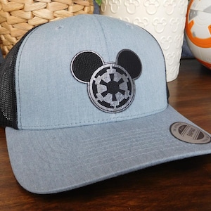 May include: A light blue trucker hat with a black mesh back and a curved brim. The hat features a black leatherette Mickey Mouse head with a silver and black Imperial symbol. A small circular tag is on the brim.