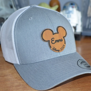 May include: A gray and white baseball cap with a brown wooden Mickey Mouse ears patch. The patch has the name "Emma" and the text "Disney 2021" on it.