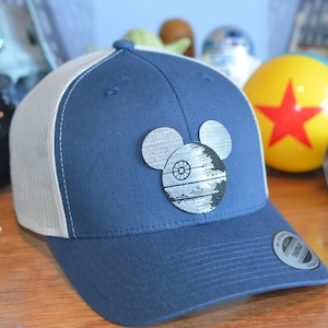 May include: A blue and white trucker hat with a black and white Mickey Mouse design featuring the Death Star. The hat has a mesh back and a snap closure.