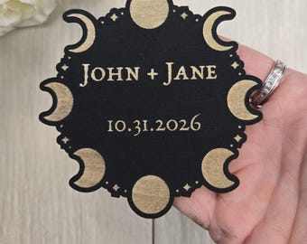 May include: A black, laser-cut wooden wedding favor with a celestial design. The text "JOHN + JANE" and the date "10.31.2026" are in a gold color. The design includes moons and stars.
