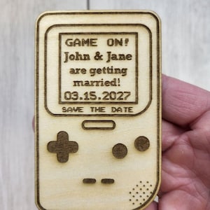 Op de afbeelding: Een houten save-the-date aankondiging in de vorm van een retro handheld gameconsole. De tekst "GAME ON! John & Jane are getting married! 03.15.2027 SAVE THE DATE" is op de voorkant gegraveerd. Het ontwerp bevat een D-pad en knoppen.