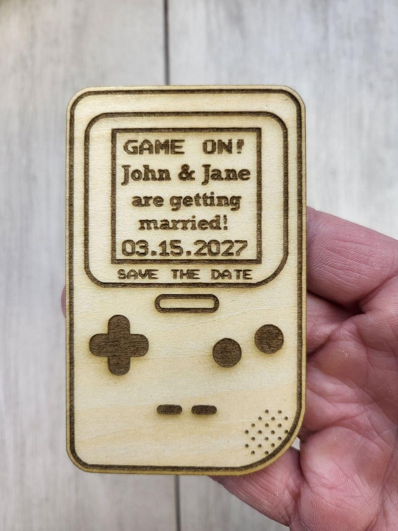 Op de afbeelding: Een houten save-the-date aankondiging in de vorm van een retro handheld gameconsole. De tekst "GAME ON! John & Jane are getting married! 03.15.2027 SAVE THE DATE" is op de voorkant gegraveerd. Het ontwerp bevat een D-pad en knoppen.