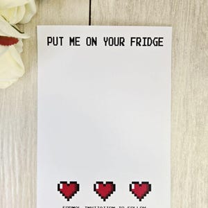 Op de afbeelding: Witte uitnodigingskaart met de tekst "PUT ME ON YOUR FRIDGE" in pixellettertype. Onderin staan drie gepixelde rode harten, met daaronder de tekst "FORMAL INVITATION TO FOLLOW". Witte bloemen op de achtergrond.