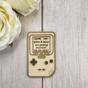 Op de afbeelding: Een houten save-the-date aankondiging in de vorm van een retro handheld game console. De tekst "GAME ON! John & Jane are getting married! 03.15.2027 SAVE THE DATE" is gegraveerd op het scherm. De aankondiging is lichtbruin.