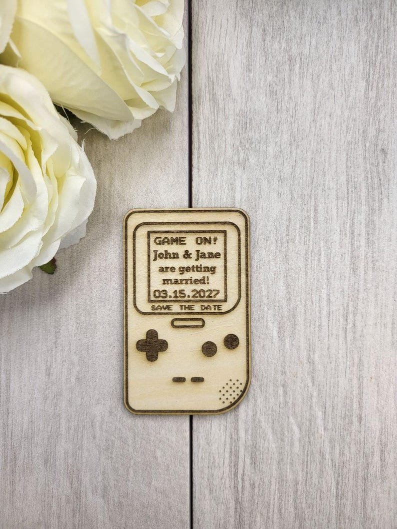 Op de afbeelding: Een houten save-the-date aankondiging in de vorm van een retro handheld game console. De tekst "GAME ON! John & Jane are getting married! 03.15.2027 SAVE THE DATE" is gegraveerd op het scherm. De aankondiging is lichtbruin.