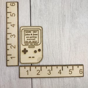 Op de afbeelding: Houten save-the-date aankondiging in de vorm van een retro handheld gameconsole. De tekst op het scherm luidt "GAME ON! John & Jane are getting married! 03.15.2027 SAVE THE DATE." Twee houten linialen zijn ook zichtbaar.