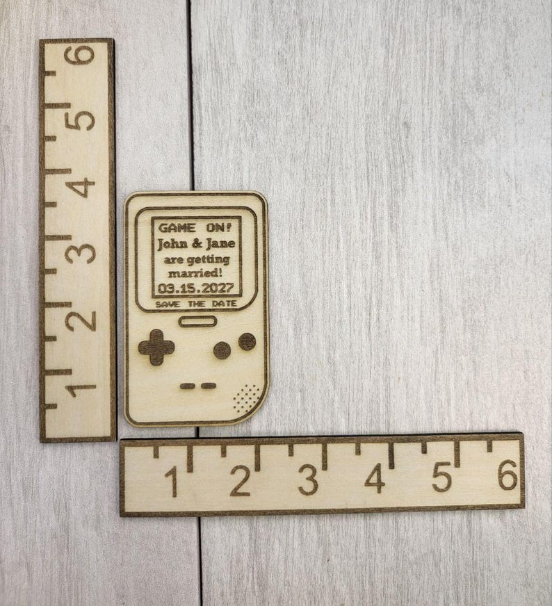 Op de afbeelding: Houten save-the-date aankondiging in de vorm van een retro handheld gameconsole. De tekst op het scherm luidt "GAME ON! John & Jane are getting married! 03.15.2027 SAVE THE DATE." Twee houten linialen zijn ook zichtbaar.