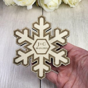 May include: A wooden snowflake ornament with a brown outline and a white center. The ornament is engraved with the initials "J+C" and the date "09.25.2025".