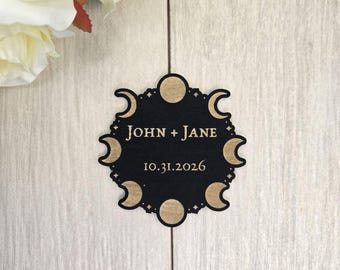 Op de afbeelding: Zwart en gouden huwelijksbedankje met een hemels ontwerp. Het cirkelvormige label toont halve manen, volle manen en de namen "JOHN + JANE" met de datum "10.31.2026".