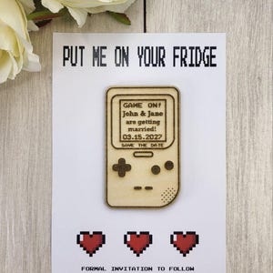 Op de afbeelding: Een save-the-date kaart met een houten gameconsole ontwerp. De kaart leest "PUT ME ON YOUR FRIDGE" en bevat de tekst "GAME ON! John & Jane are getting married! 03.15.2027 SAVE THE DATE." Hieronder staan drie gepixelde rode harten en de tekst "FORMAL INVITATION TO FOLLOW."