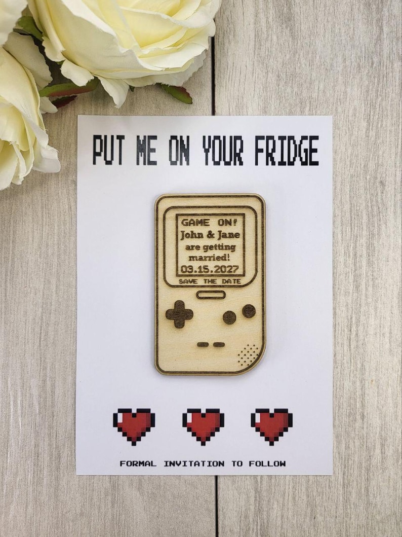 Op de afbeelding: Een save-the-date kaart met een houten gameconsole ontwerp. De kaart leest "PUT ME ON YOUR FRIDGE" en bevat de tekst "GAME ON! John & Jane are getting married! 03.15.2027 SAVE THE DATE." Hieronder staan drie gepixelde rode harten en de tekst "FORMAL INVITATION TO FOLLOW."