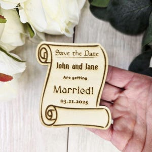Könnte beinhalten: Holz-Save-the-Date-Ankündigung in Form einer Schriftrolle. Die Schriftrolle ist mit dem Text "Save the Date", "John and Jane", "Are getting Married!" und "03.21.2025" graviert.