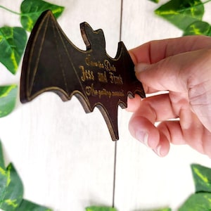 May include: A wooden bat-shaped save the date card with the text "Save the Date Jess and Frank We're getting married" in gold lettering.