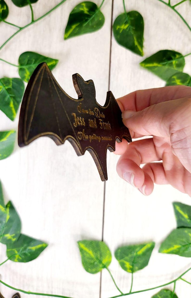 May include: A wooden bat-shaped save the date card with the text "Save the Date Jess and Frank We're getting married" in gold lettering.