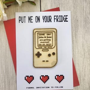 Op de afbeelding: Een trouwkaart met een retro videogame thema. De kaart heeft een houten gameconsole ontwerp met de tekst "GAME ON!" en "SAVE THE DATE". De kaart bevat ook de tekst "PUT ME ON YOUR FRIDGE" en "FORMAL INVITATION TO FOLLOW".