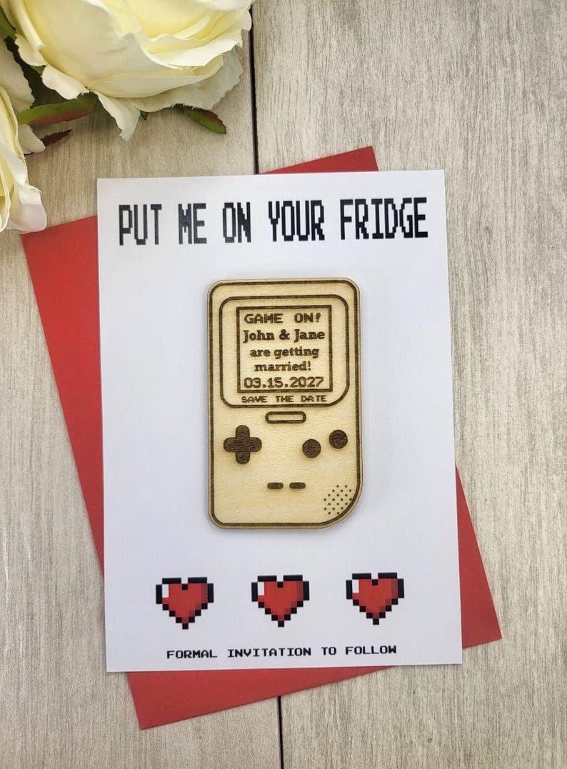 Op de afbeelding: Een trouwkaart met een retro videogame thema. De kaart heeft een houten gameconsole ontwerp met de tekst "GAME ON!" en "SAVE THE DATE". De kaart bevat ook de tekst "PUT ME ON YOUR FRIDGE" en "FORMAL INVITATION TO FOLLOW".
