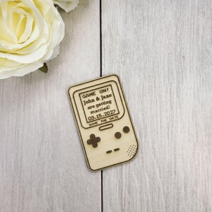 Op de afbeelding: Een houten save-the-date aankondiging in de vorm van een retro handheld gameconsole. De tekst "GAME ON! John & Jane are getting married! 03.15.2027 SAVE THE DATE" is gegraveerd op het scherm. De achtergrond is een licht houten oppervlak.