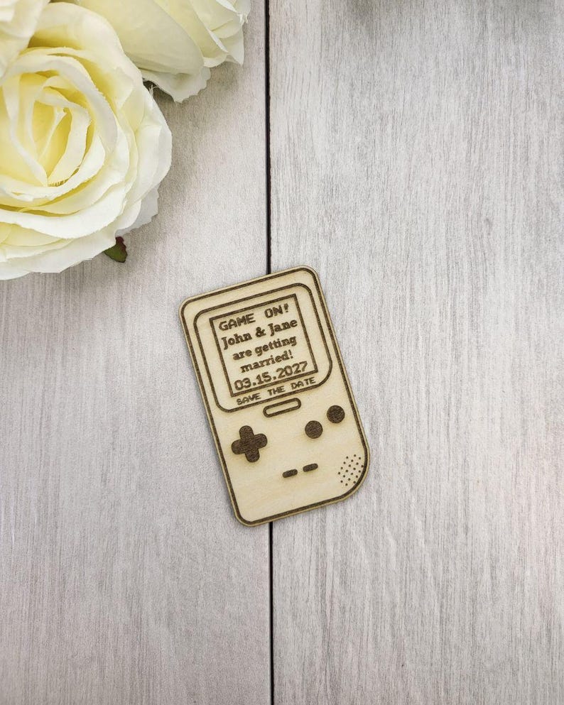 Op de afbeelding: Een houten save-the-date aankondiging in de vorm van een retro handheld gameconsole. De tekst "GAME ON! John & Jane are getting married! 03.15.2027 SAVE THE DATE" is gegraveerd op het scherm. De achtergrond is een licht houten oppervlak.