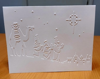 May include: A white embossed Christmas card featuring the three wise men, a camel, a star, and a cityscape. The card is made of thick paper and has a simple, elegant design. The embossed details add a tactile element.