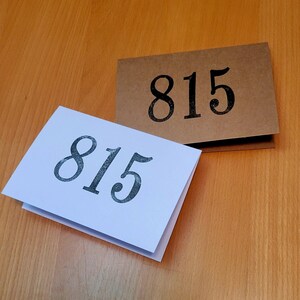 May include: Two folded cards with the number "815" printed on them. One card is white with dark gray text, and the other is brown with black text. The cards are on a wooden surface.