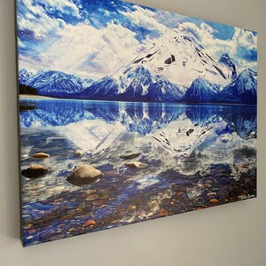 May include: A canvas print of a snowy mountain range reflected in a still lake. The water is clear and blue, with small rocks visible at the bottom. The sky is a bright blue with white clouds. The mountains are covered in snow and ice. The print is signed "T. Haas Butler" in the lower right corner.