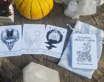 May include: Three oracle cards with black and white illustrations on a white background. The cards are laid out on a wooden surface with a small pumpkin, a crystal cluster, and a white crystal. The cards read "Turn the Page", "Pay Attention", and "Trust the Process". The bag of cards reads "Intuitive Oracle Cards" and "Melanie Beckler".