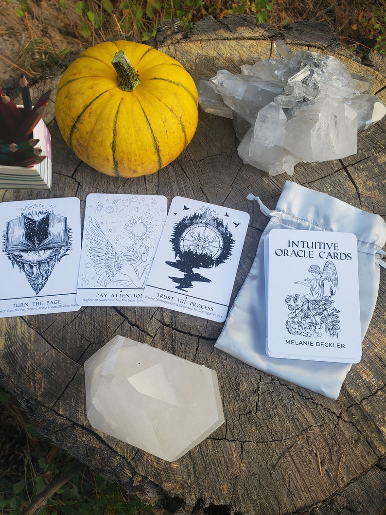 May include: Three oracle cards with black and white illustrations on a white background. The cards are laid out on a wooden surface with a small pumpkin, a crystal cluster, and a white crystal. The cards read "Turn the Page", "Pay Attention", and "Trust the Process". The bag of cards reads "Intuitive Oracle Cards" and "Melanie Beckler".