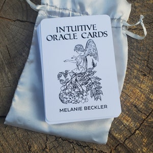 May include: A white deck of Intuitive Oracle Cards with a black and white illustration of an angel sitting on a branch with berries. The text on the card reads "Intuitive Oracle Cards" and "Melanie Beckler".