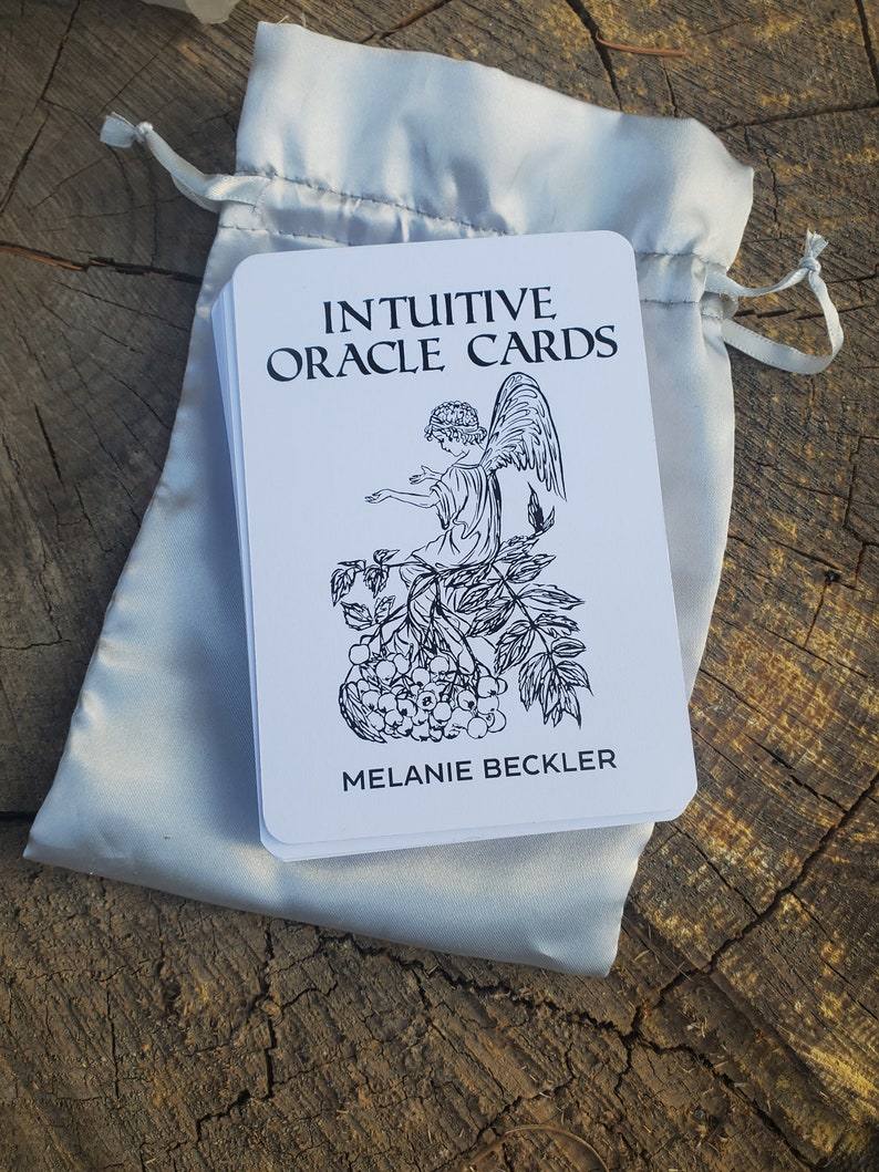 May include: A white deck of Intuitive Oracle Cards with a black and white illustration of an angel sitting on a branch with berries. The text on the card reads "Intuitive Oracle Cards" and "Melanie Beckler".