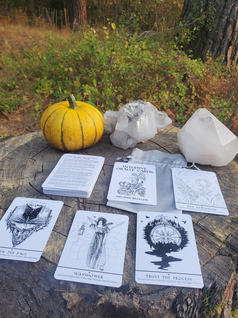 May include: A set of intuitive oracle cards with illustrations of a book, a woman with wings, a compass, and a bird. The cards are laid out on a wooden surface with a small pumpkin, crystals, and a white cloth.