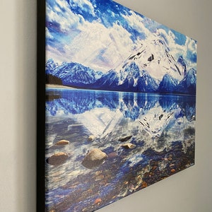May include: A canvas print of a snowy mountain range reflected in a still lake. The sky is a vibrant blue with fluffy white clouds. The water is clear and blue, with rocks visible at the bottom.
