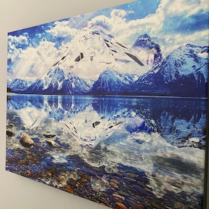 May include: A painting of a snowy mountain range reflected in a still lake. The mountain peaks are white and the sky is blue with white clouds. The lake is a deep blue and the water is calm. The painting is signed "Melanie Beckler" in the lower right corner.