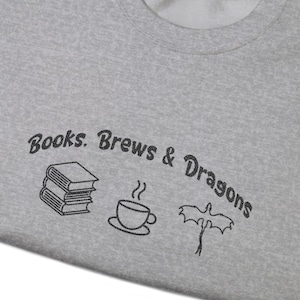 May include: A grey sweatshirt featuring the words "Books, Brews & Dragons" and illustrations of a stack of books, a coffee cup, and a dragon. The sweatshirt is folded, displaying the design.