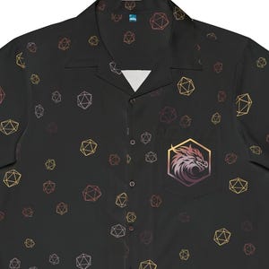 Op de afbeelding: Zwart overhemd met knopen en een herhalend patroon van kleurrijke polyhedrale dobbelstenen. Een zak op de linkerborst heeft een drakenembleem in een gouden zeshoek. Het shirt met korte mouwen heeft een kampkraag.