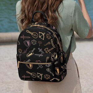 Könnte beinhalten: Schwarzer Rucksack mit goldenem Reißverschluss und Trägern. Der Rucksack hat ein sich wiederholendes Muster von Fantasy-Symbolen in Gold, Rosa und Weiß. Die Person im Bild trägt einen Strohhut mit schwarzem Band und beige Shorts.
