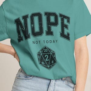May include: Teal t-shirt with the word "NOPE" in distressed black lettering. Below the word "NOPE" is the phrase "NOT TODAY" and a graphic of a 20-sided die. The model is wearing denim shorts.