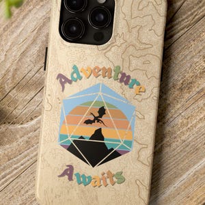 May include: A beige phone case with a colorful graphic of a 20-sided die with a dragon flying over a mountain range. The text "Adventure Awaits" is printed on the case.