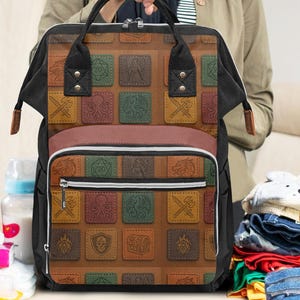 May include: A black and brown diaper bag with a decorative pattern of colorful squares. The bag has a top handle and a front zipper pocket. Baby clothes and shoes are next to the bag.