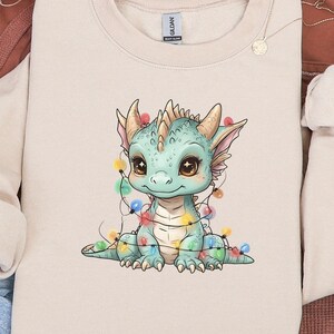 Può includere: Una felpa girocollo beige con un drago dei cartoni animati con squame blu e verdi, adornato con luci natalizie colorate. Il drago ha grandi occhi marroni ed è seduto. La felpa fa parte di un set di abbigliamento casual.