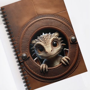 May include: A brown leather notebook with a spiral binding. A small, white dragon with large eyes peeks out from a circular opening in the cover.