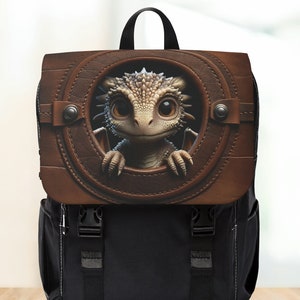Peut inclure: Un sac à dos noir avec un rabat en cuir marron sur lequel un dragon de dessin animé sort d'une ouverture circulaire. Le dragon est blanc et gris avec de grands yeux.